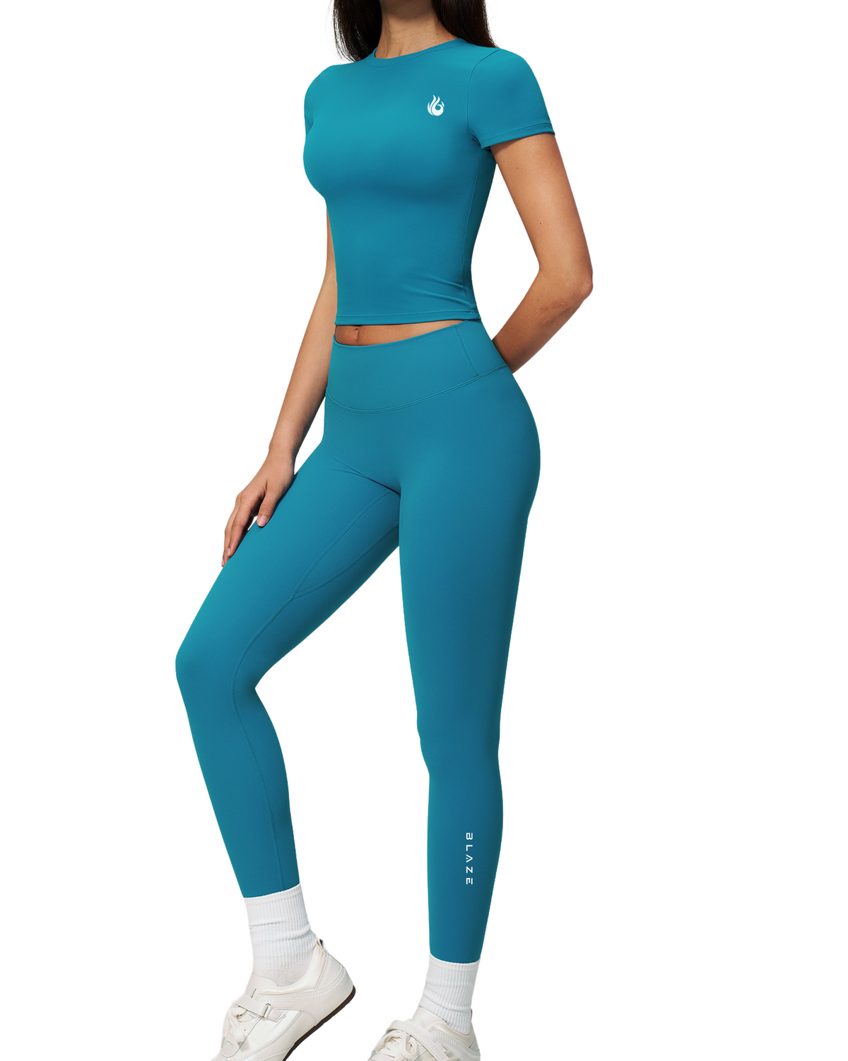 A woman modeling a matching teal blue activewear set consisting of a short-sleeve crop top and high-waisted leggings, paired with white socks and white sneakers, set against a plain white background.